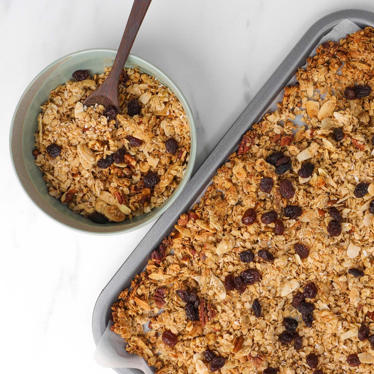 Granola A bowl of granola with a wooden spoon next to a tray of baked granola.