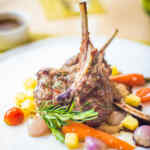 Daging domba panggang disajikan dengan sayur-sayuran berwarna dan rempah-rempah di atas piring putih.