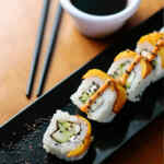 Un primer plano de rollos de sushi con semillas de sésamo naranja y negro en un plato negro, acompañados de salsa de soja.