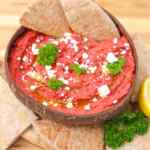 Un tazón de hummus de remolacha vibrante adornado con hierbas y servido con chips de pita.