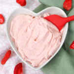 Mangkuk berbentuk hati berisi krim keju stroberi merah muda dan spatula merah.