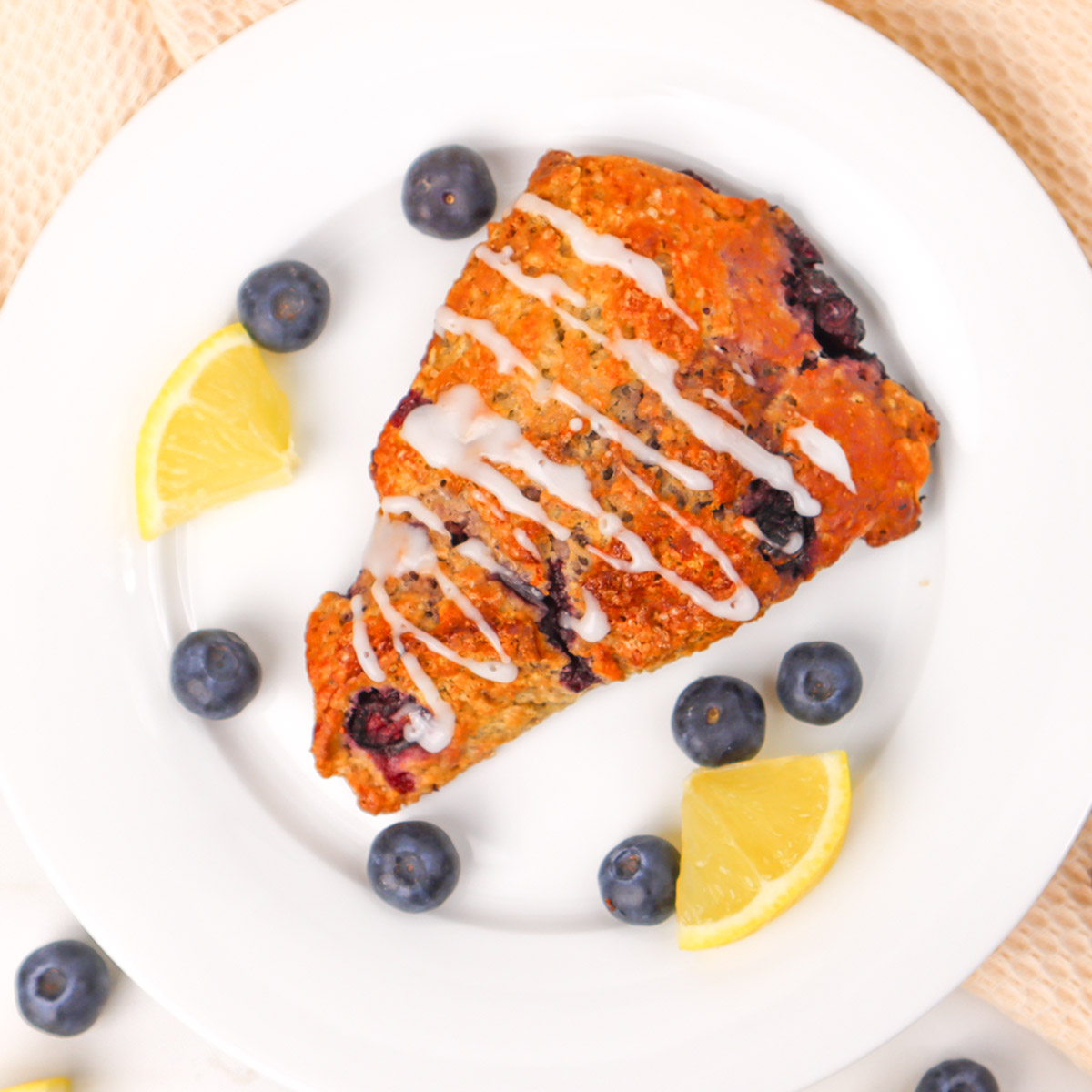 Ein mit Zuckerguss beträufelter Blaubeer-Scone, serviert mit frischen Blaubeeren und Zitronenspalten auf einem weißen Teller.