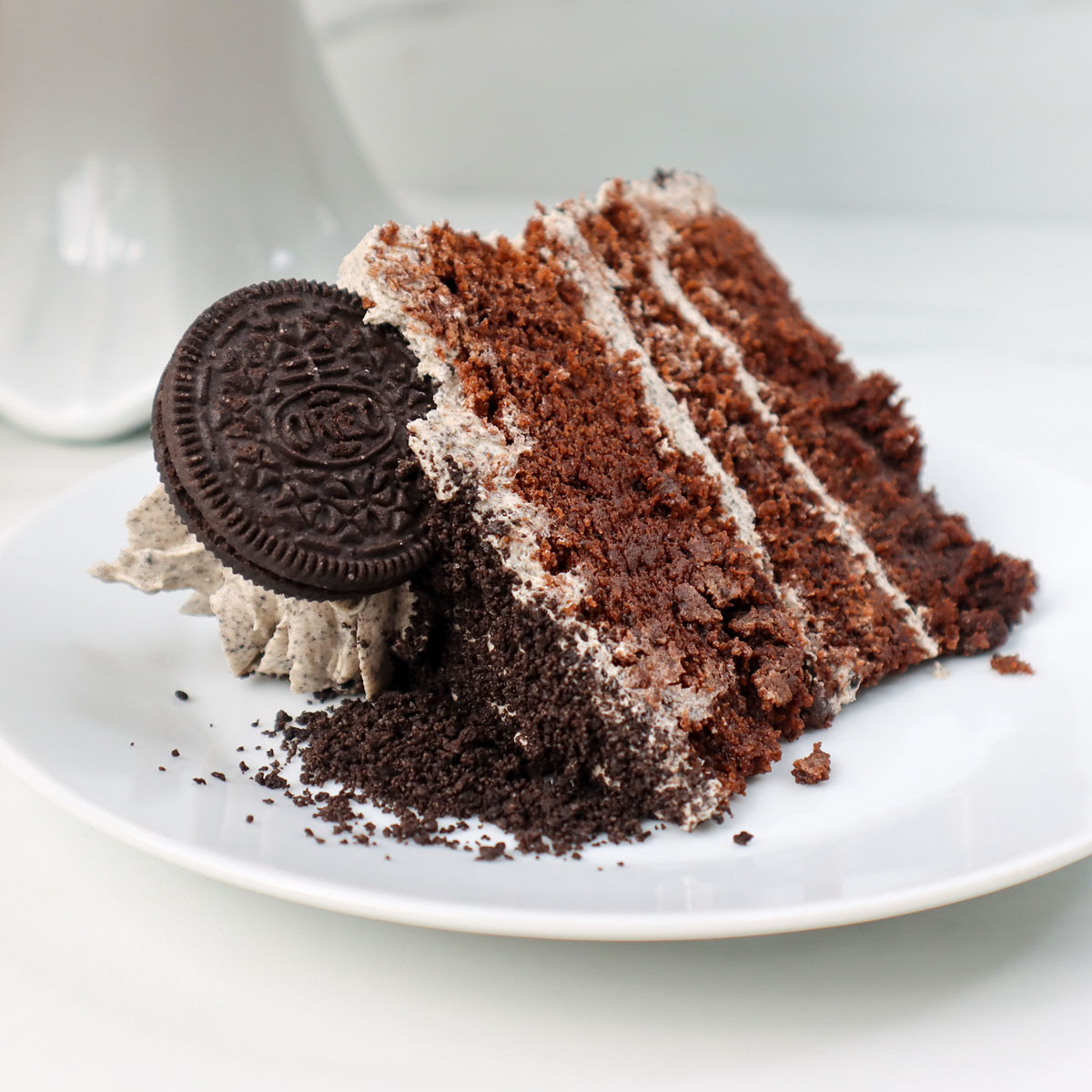 Sepotong kue coklat dengan krim lapisan dan remah kue di atas piring putih.