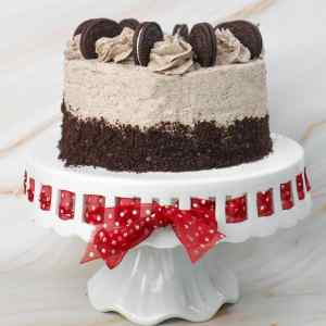 Kue Oreo berlapis-lapis, diberi potongan kue Oreo dan gulungan frosting di atas tatakan kue dekoratif.
