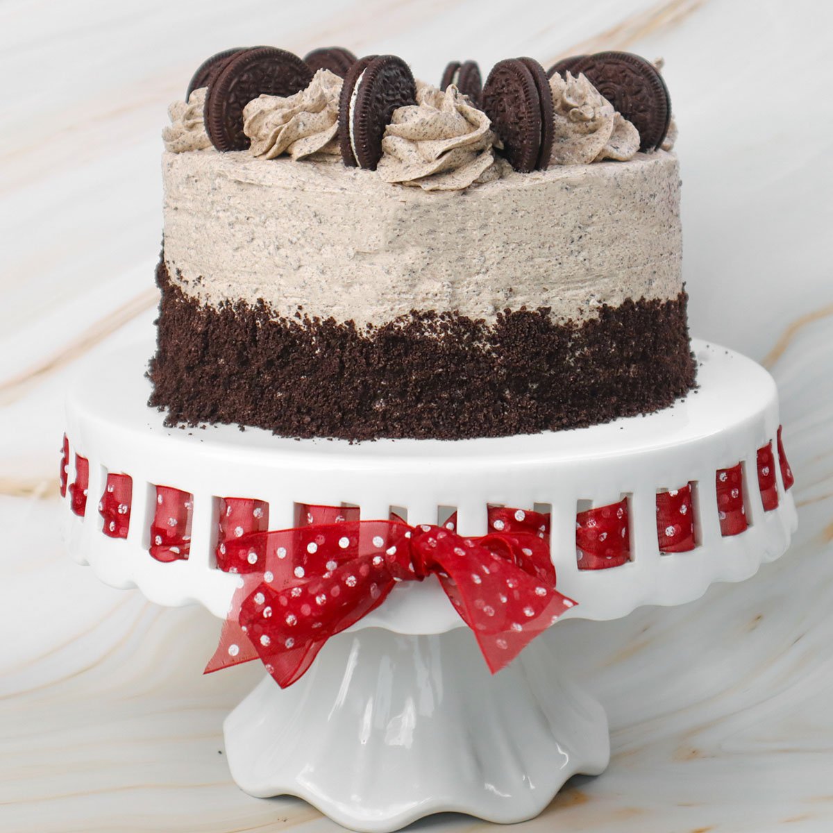 Kue Oreo berlapis-lapis, diberi potongan kue Oreo dan gulungan frosting di atas tatakan kue dekoratif.