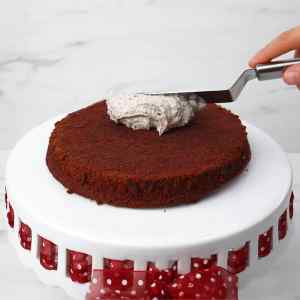 Sebuah tangan mengoleskan lapisan Oreo pada kue coklat di atas tatakan kue putih.