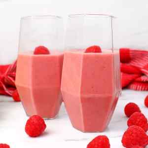 Deux verres remplis de smoothie framboise-banane surmontés d'une framboise fraîche, entourés de framboises fraîches et d'un torchon rouge.