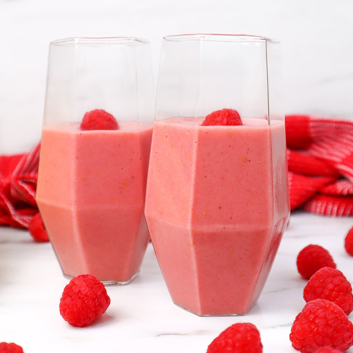Deux verres remplis de smoothie framboise-banane surmontés d'une framboise fraîche, entourés de framboises fraîches et d'un torchon rouge.