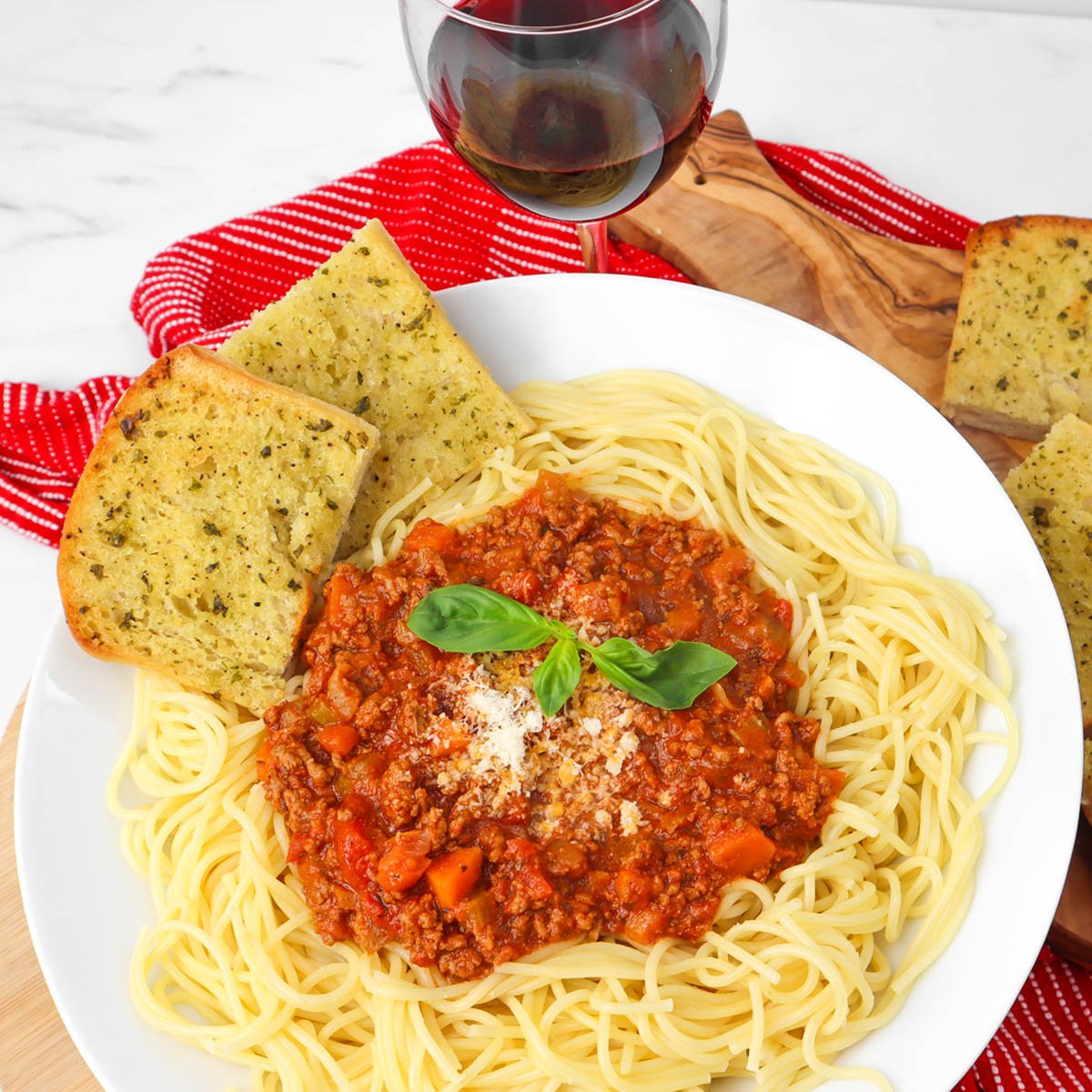 A plate of spaghetti Bolognese topped with basil and served with garlic bread and a glass of red wine.