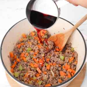 Red wine is being poured into a pot of sautéed ground meat and vegetables for spaghetti Bolognese.
