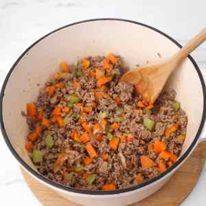 Cooked ground meat mixed with diced carrots, celery, and onions in a pot.