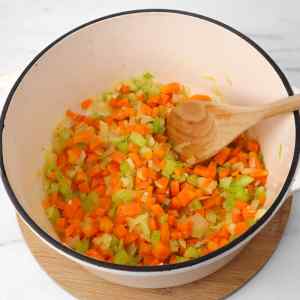 Chopped carrots, celery, and onions sautéing in a pot with a wooden spoon.