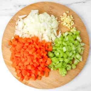 Chopped onions, carrots, celery, and garlic arranged on a wooden cutting board.
