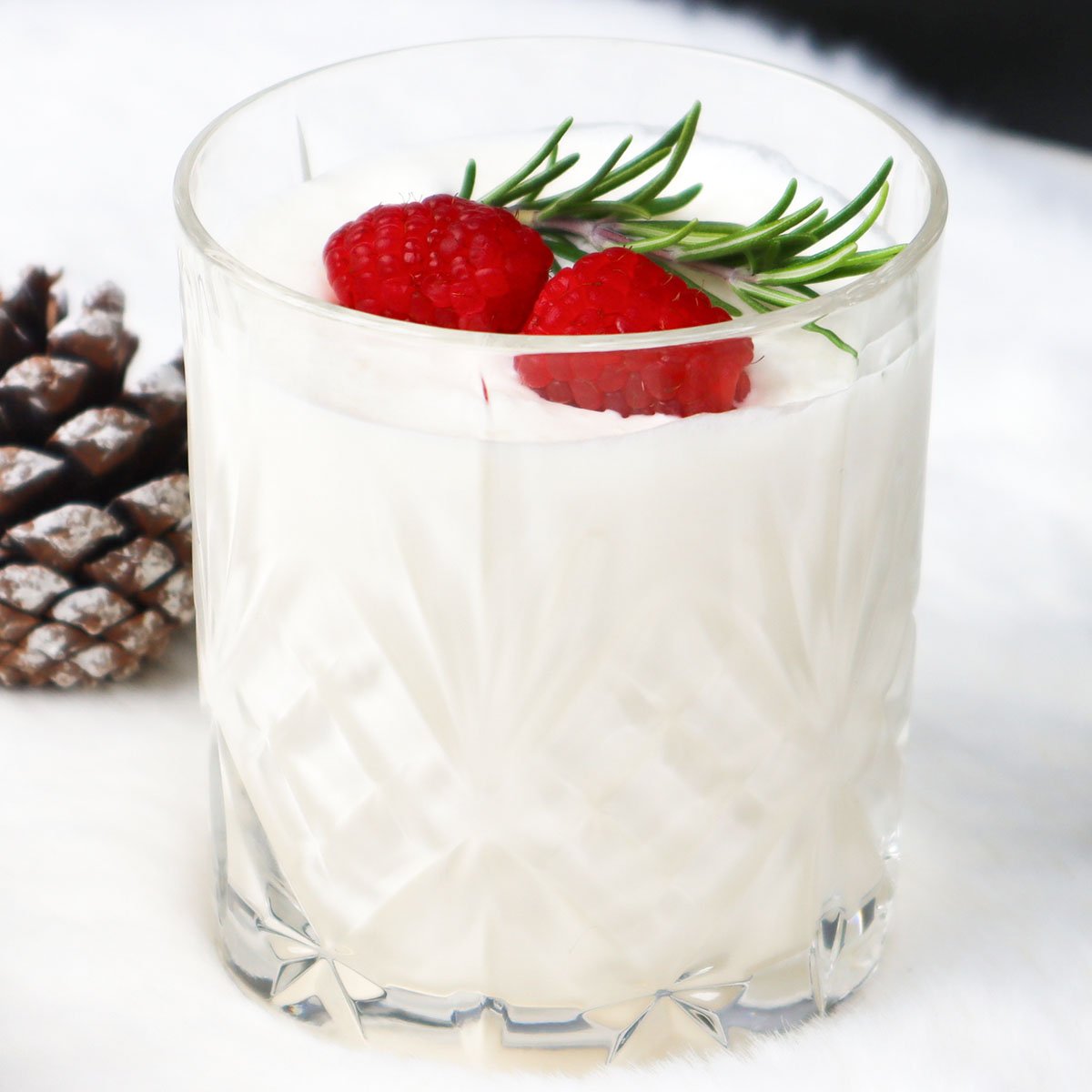 Receita de coquetel de Natal branco 1200-03 Um coquetel branco cremoso decorado com framboesas e alecrim, sobre um fundo suave.