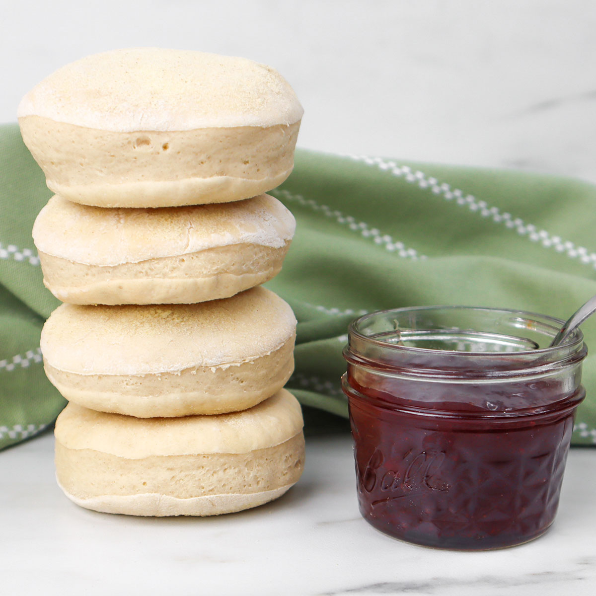 Quatre muffins anglais cuits à la friteuse à air, empilés à côté d'un pot de confiture sur une surface en marbre.