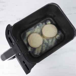 Deux muffins anglais crus reposent dans le panier d'une friteuse à air, saupoudrés de semoule.