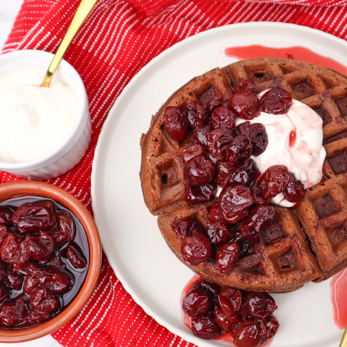 Chocolate waffles topped with cherries and cream on a red striped cloth.