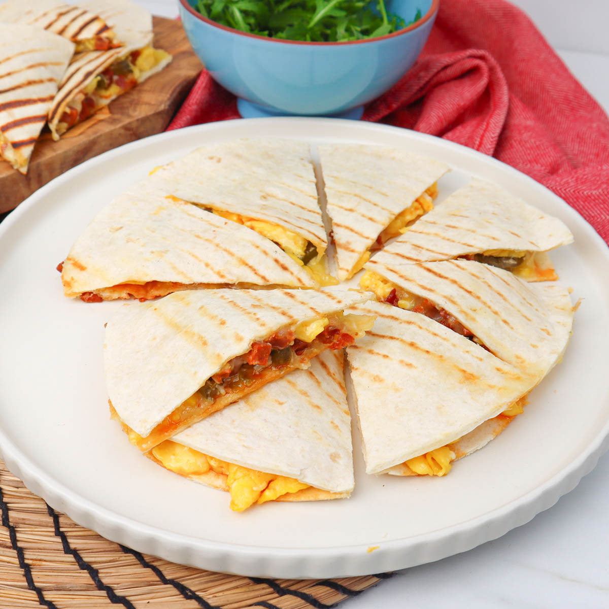 Quesadillas du petit-déjeuner Des quesadillas coupées en tranches, garnies d'œufs et de légumes, sont présentées dans une assiette, avec un bol de salade verte en arrière-plan.