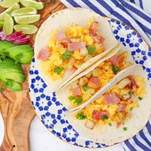 Trois tacos pour le petit-déjeuner garnis d'œufs brouillés, de jambon et de pommes de terre, agrémentés de coriandre et présentés sur une assiette décorative.