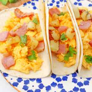 Trois tacos pour le petit-déjeuner garnis d'œufs brouillés, de jambon et de pommes de terre en dés, le tout agrémenté de coriandre.