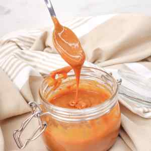 A spoon drips caramel sauce from a glass jar, set against a soft fabric background.