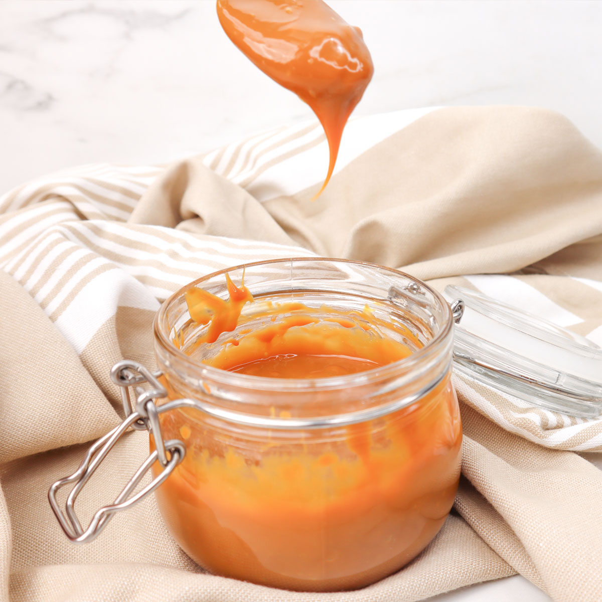 Caramel Sauce A jar of caramel sauce with a spoon dripping caramel, set on a light fabric background.
