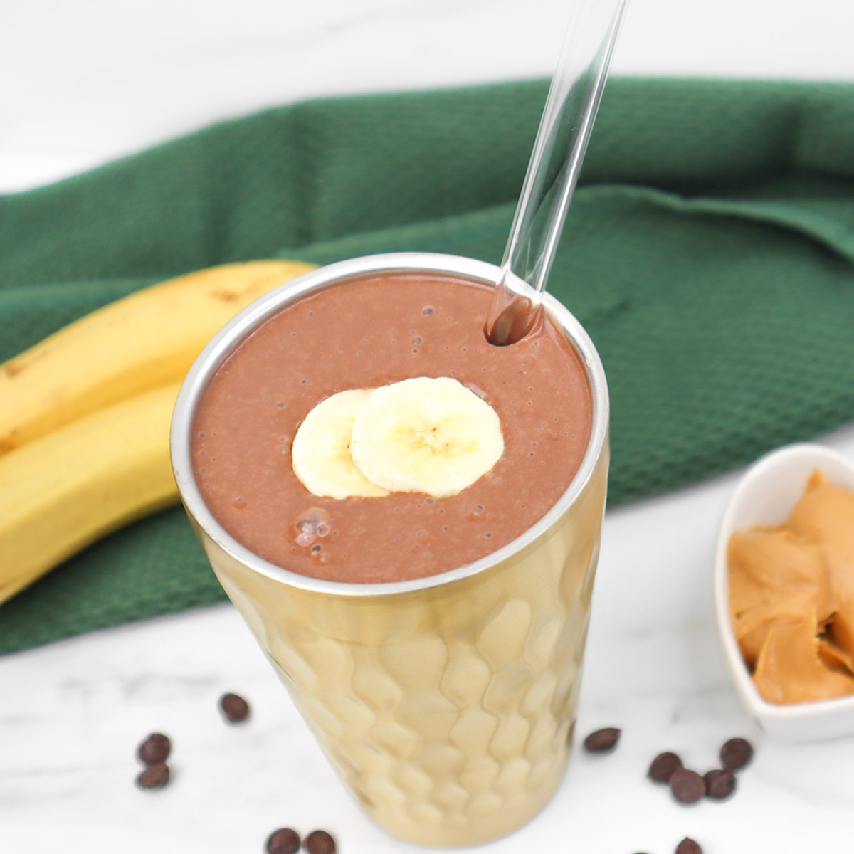 A chocolate peanut butter smoothie topped with banana slices, next to bananas and peanut butter.