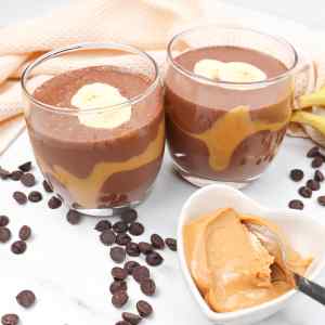 Deux verres de smoothie au chocolat et au beurre de cacahuète, garnis de tranches de banane, le tout entouré de pépites de chocolat et de beurre de cacahuète.