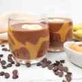 Twee glazen chocolade-pindakaassmoothie, gegarneerd met plakjes banaan, omringd door chocoladeschilfers en pindakaas.