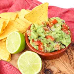 Un bol de guacamole avec des tomates en dés, servi avec des chips de tortilla et des tranches de citron vert.