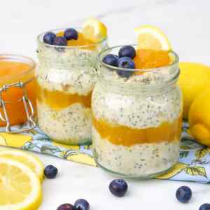 Dois potes de aveia adormecida com creme de limão, cobertos com mirtilos e fatias de limão, com limões e um pote de creme de limão por perto.