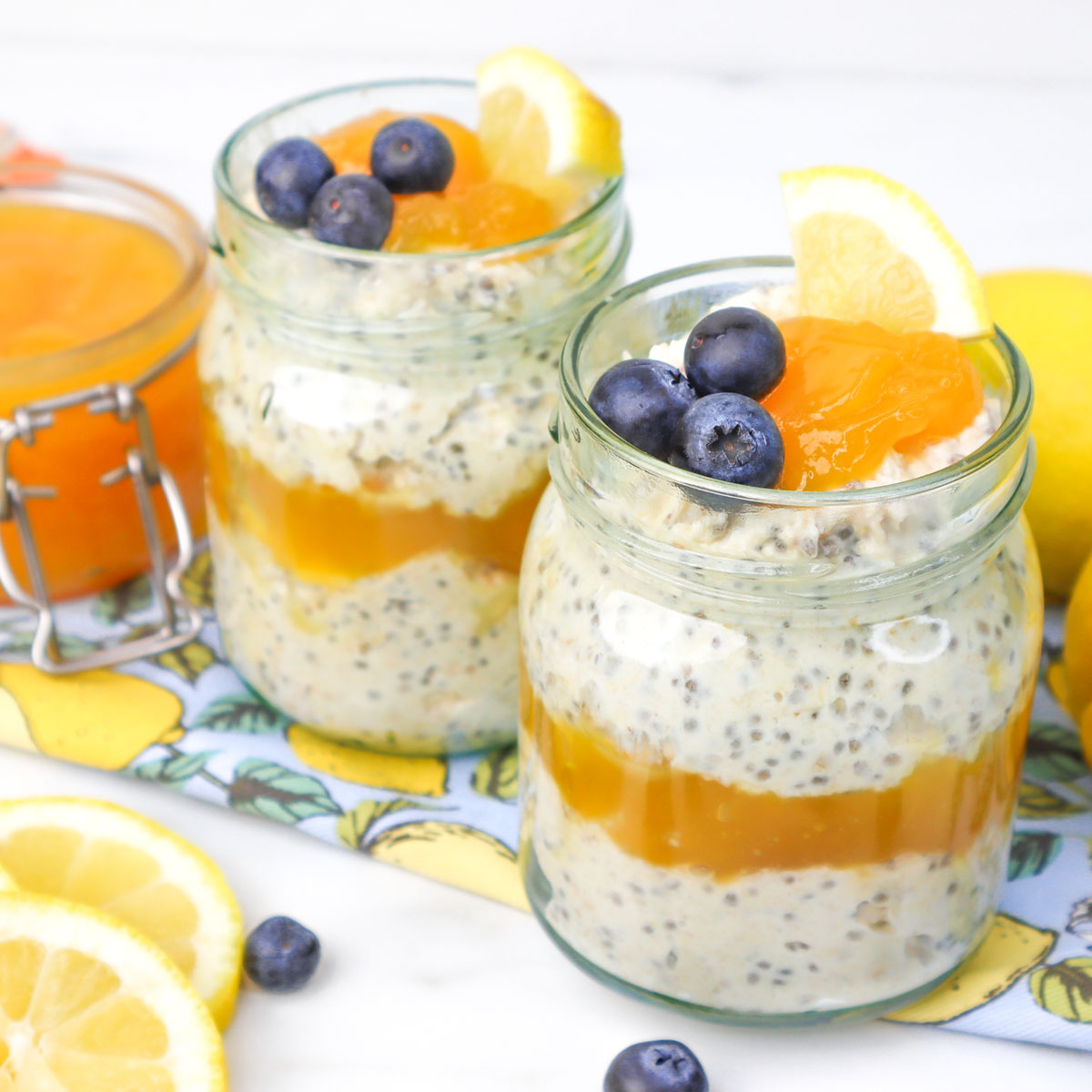 Two jars of lemon curd overnight oats topped with blueberries and lemon slices, with a jar of lemon curd nearby.