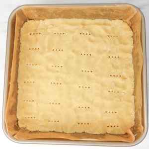 Uma forma quadrada com massa de biscoito amanteigado, marcada com pequenos furos, pronta para assar.