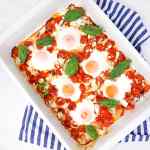 Huevos al horno con tomates y queso feta cubiertos con albahaca fresca en un plato blanco.