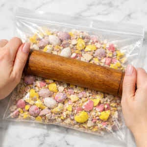 Hands crushing Mini Eggs in a freezer bag with a rolling pin for baking on a white marble countertop
