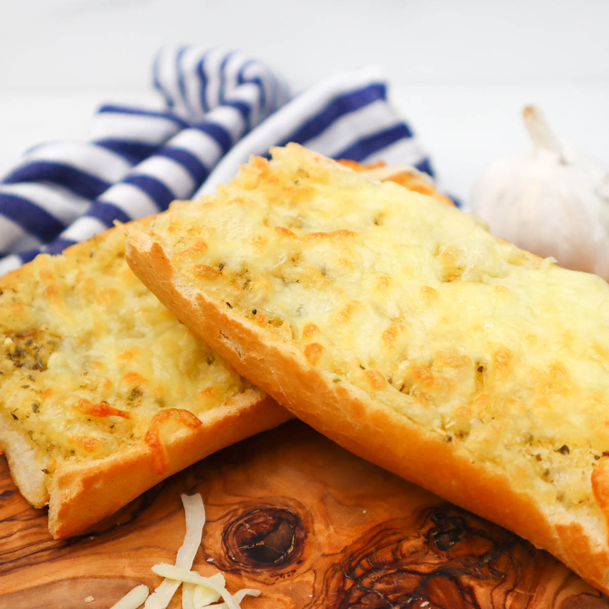 Air Fryer Cheesy Garlic Bread Two pieces of cheesy garlic bread on a wooden board, with a garlic bulb and striped cloth in the background.