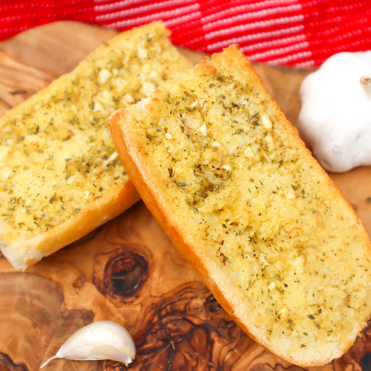 Zwei mit Kräutern bestreute Scheiben Knoblauchbrot auf einem Holzbrett, daneben eine Knoblauchzehe.