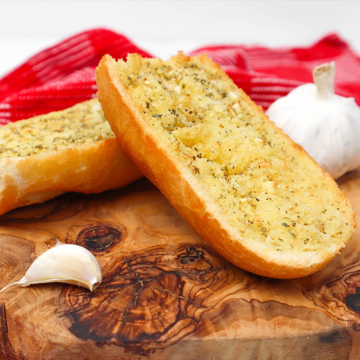 Knoblauchbrot aus der Heißluftfritteuse In Scheiben geschnittenes Knoblauchbrot, bestreut mit Kräutern, liegt auf einem Holzbrett, daneben eine Knoblauchzehe.