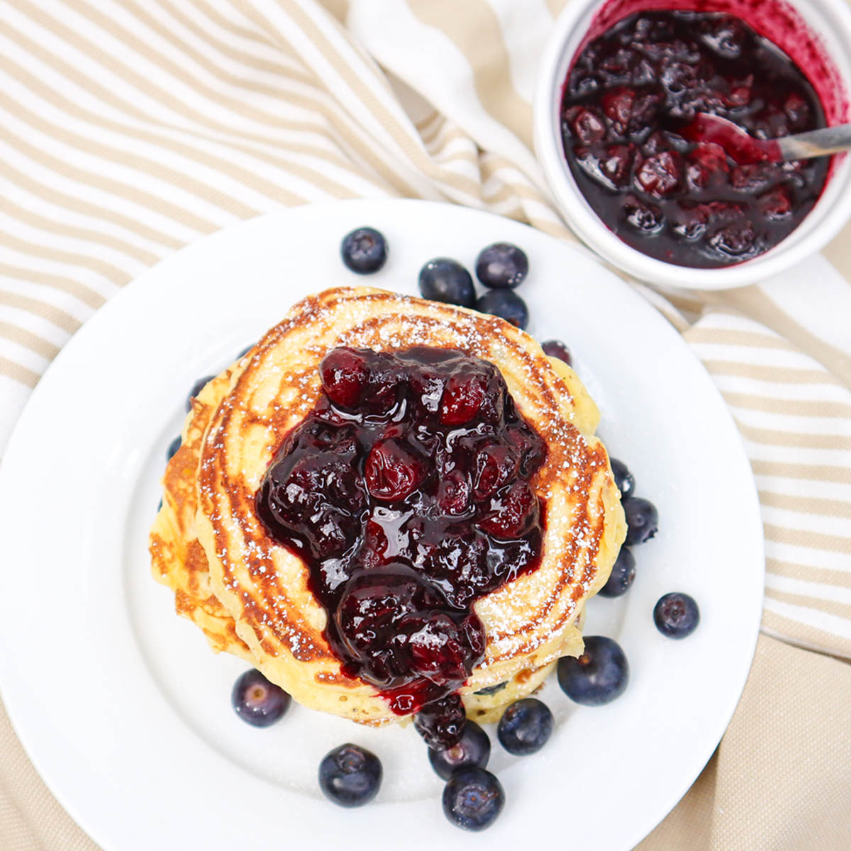 Pancake Buttermilk Blueberry Setumpuk panekuk buttermilk blueberry dengan saus beri dan blueberry segar di atas piring putih.