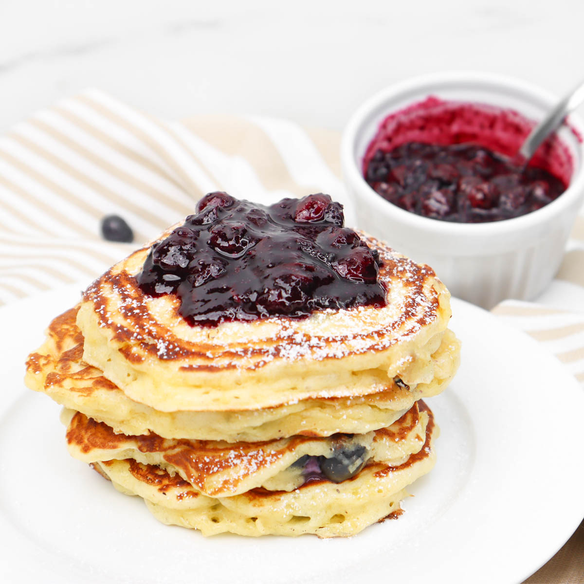 Pancake Buttermilk Blueberry Setumpuk panekuk buttermilk blueberry yang diberi saus blueberry dan gula bubuk.