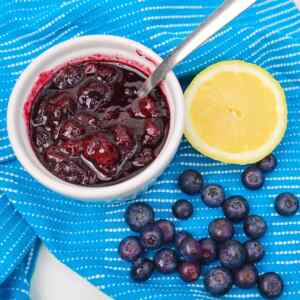 A bowl of blueberry compote with a spoon, lemon slice, and fresh blueberries on a blue patterned cloth.