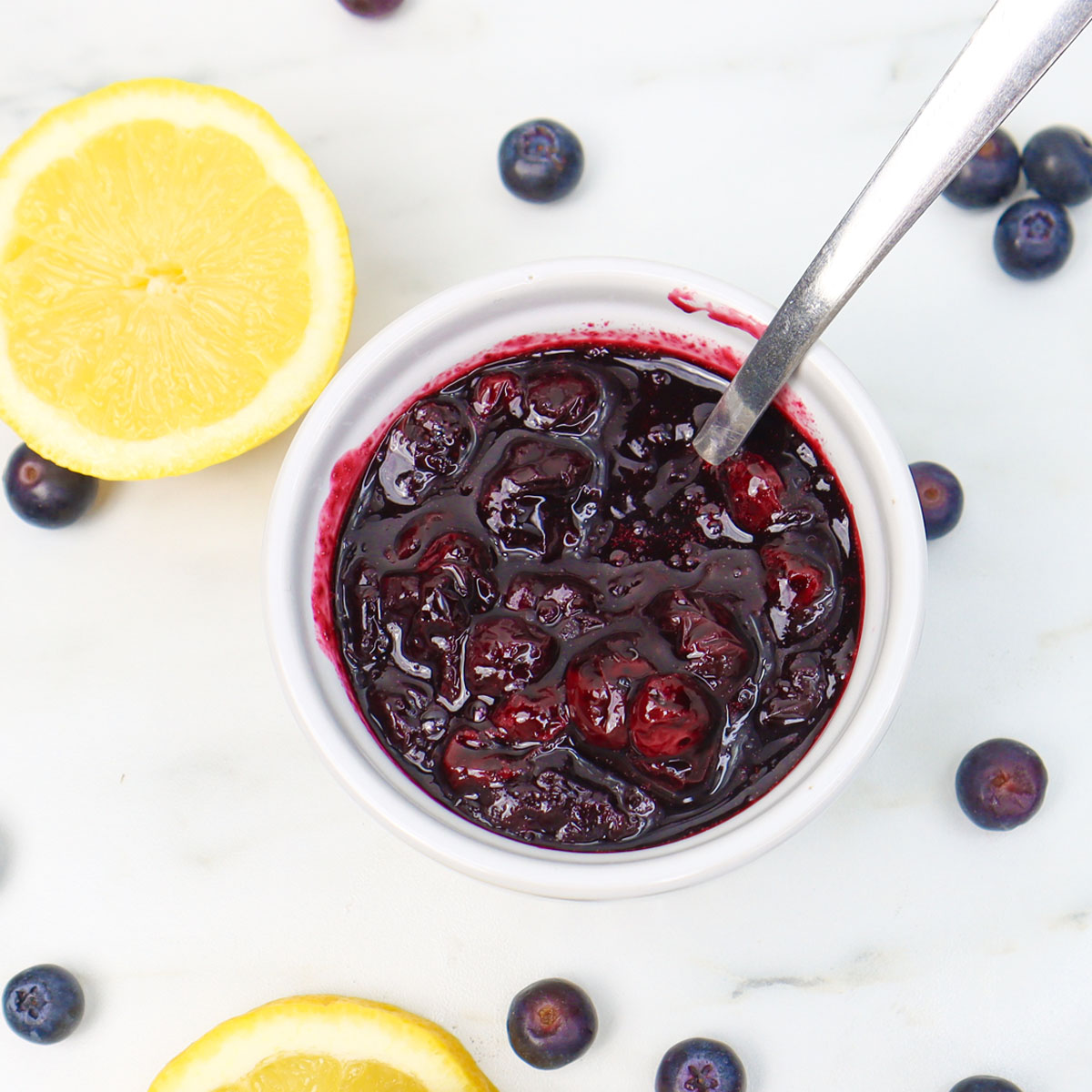 Blueberry Compote A bowl of blueberry compote with a spoon, surrounded by fresh blueberries and lemon slices.