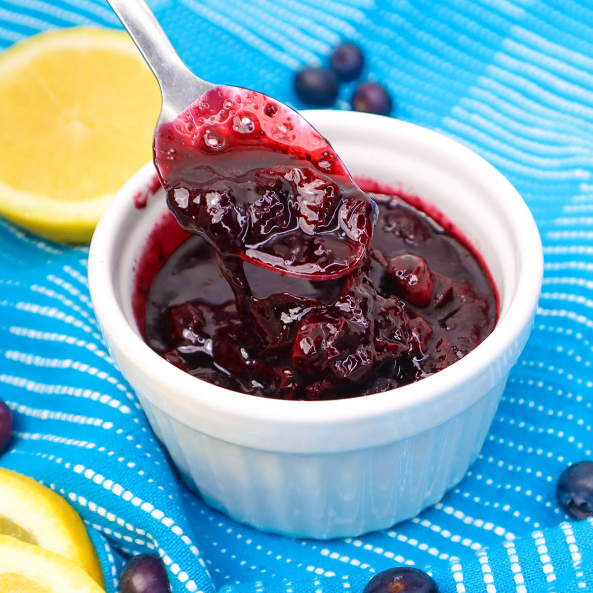 Blueberry Compote A spoonful of blueberry compote in a white dish, surrounded by fresh blueberries and lemon slices.