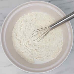 Uma tigela com farinha, açúcar, fermento em pó e sal, com um batedor de arame dentro, pronta para o preparo de panquecas.
