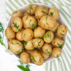 Buttery boiled new potatoes in a bowl next to a striped tea towel, and herbs scattered around on the potatoes