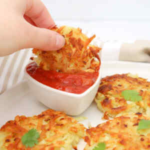 A hand dipping a crispy hash brown into a small bowl of red sauce, with more hash browns on the plate.