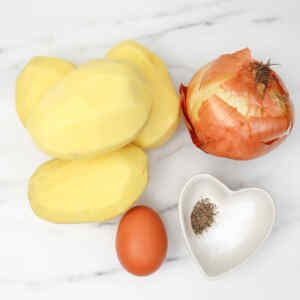 Peeled potatoes, an onion, an egg, and a small dish of seasoning on a marble surface.