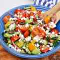 A colorful Mediterranean salad with tomatoes, cucumbers, peppers, olives, and feta cheese in a blue bowl.