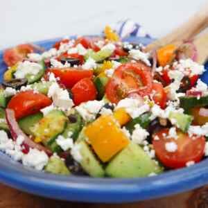 A colorful Mediterranean salad with tomatoes, cucumbers, peppers, olives, and feta cheese in a blue bowl.