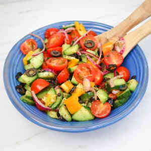 A colorful Mediterranean salad with tomatoes, cucumbers, bell peppers, olives, and red onions in a blue bowl.